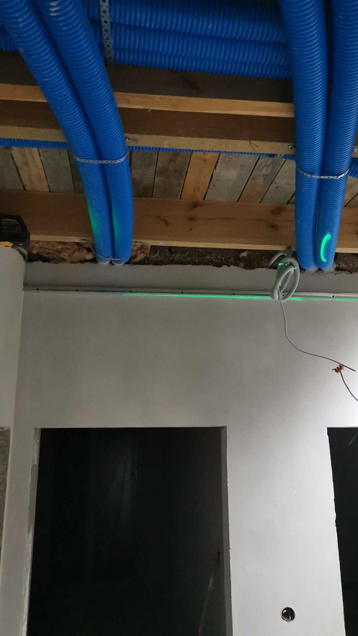 Niebieskie rury wentylacyjne pod sufitem, mocowane do drewnianej konstrukcji, z widoczną linią lasera poziomującego na ścianie. Prace wykończeniowe w trakcie.