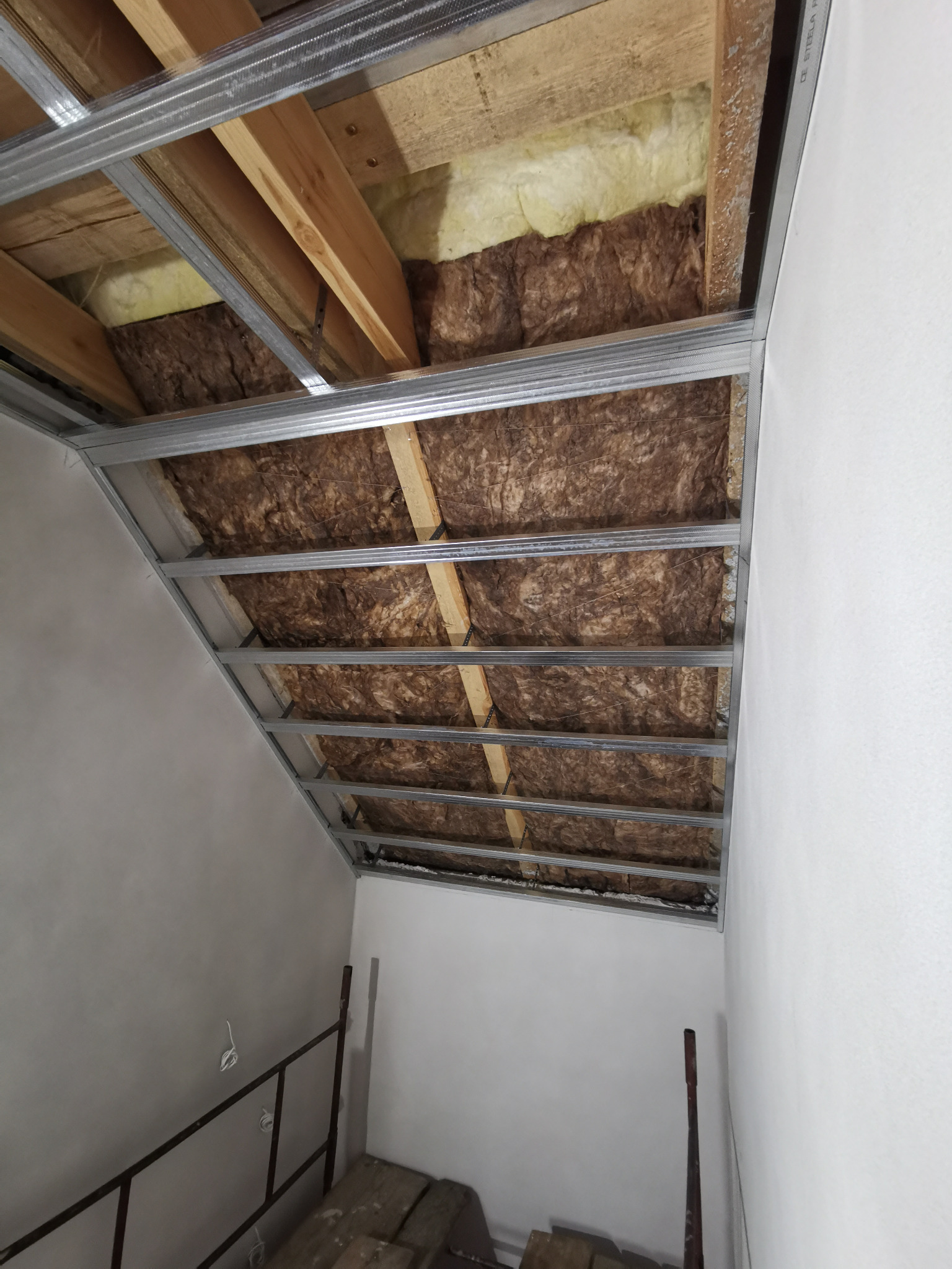 Metalowy stelaż sufitu podwieszanego z izolacją z wełny mineralnej, widok z dołu, przygotowanie do montażu płyt, ściany pomalowane na biało.