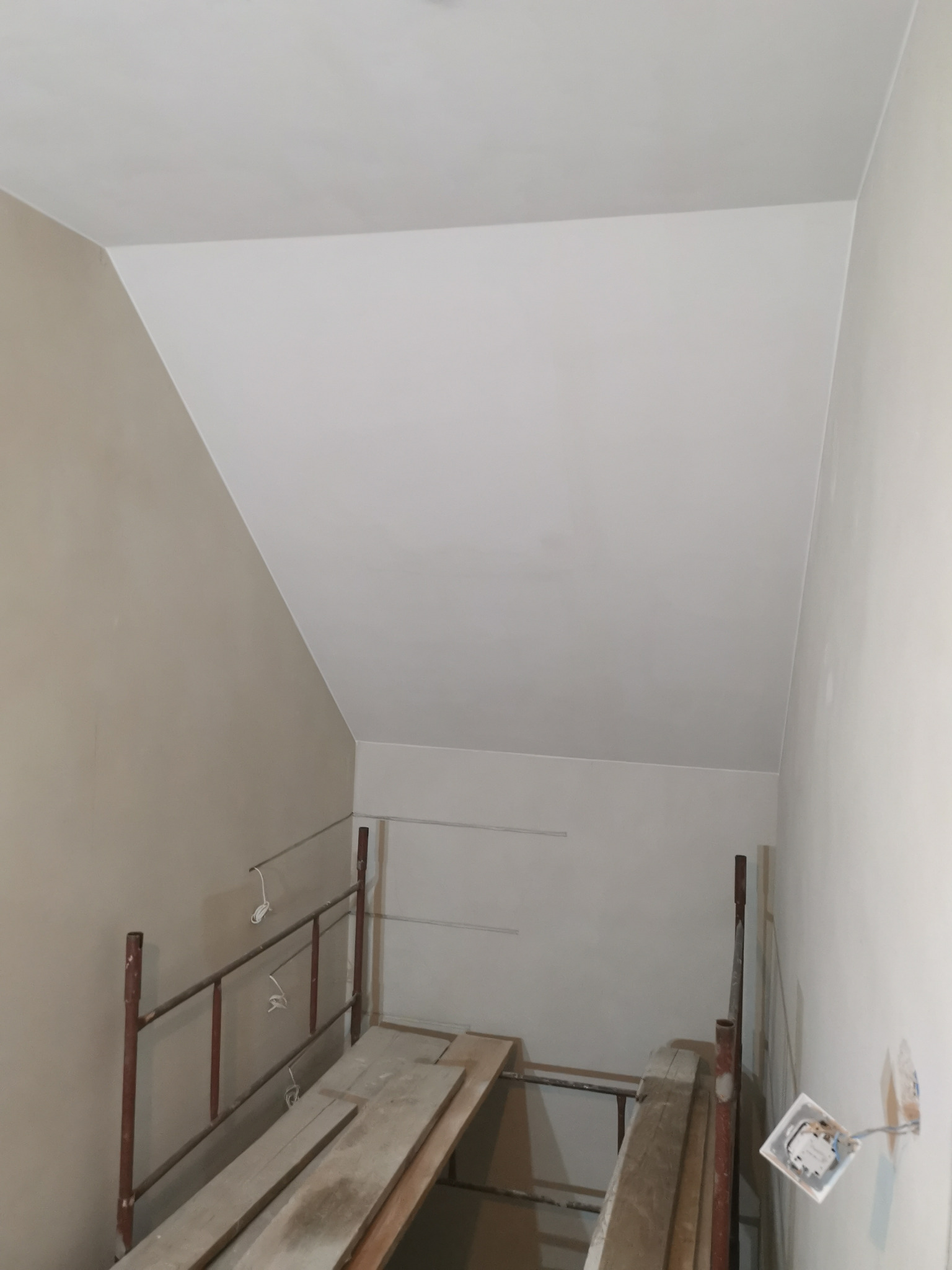 Poddasze w trakcie prac wykończeniowych: szpachlowana ściana w odcieniach beżu, rusztowanie z deskami i widoczna puszka elektryczna. Skos dachu.
