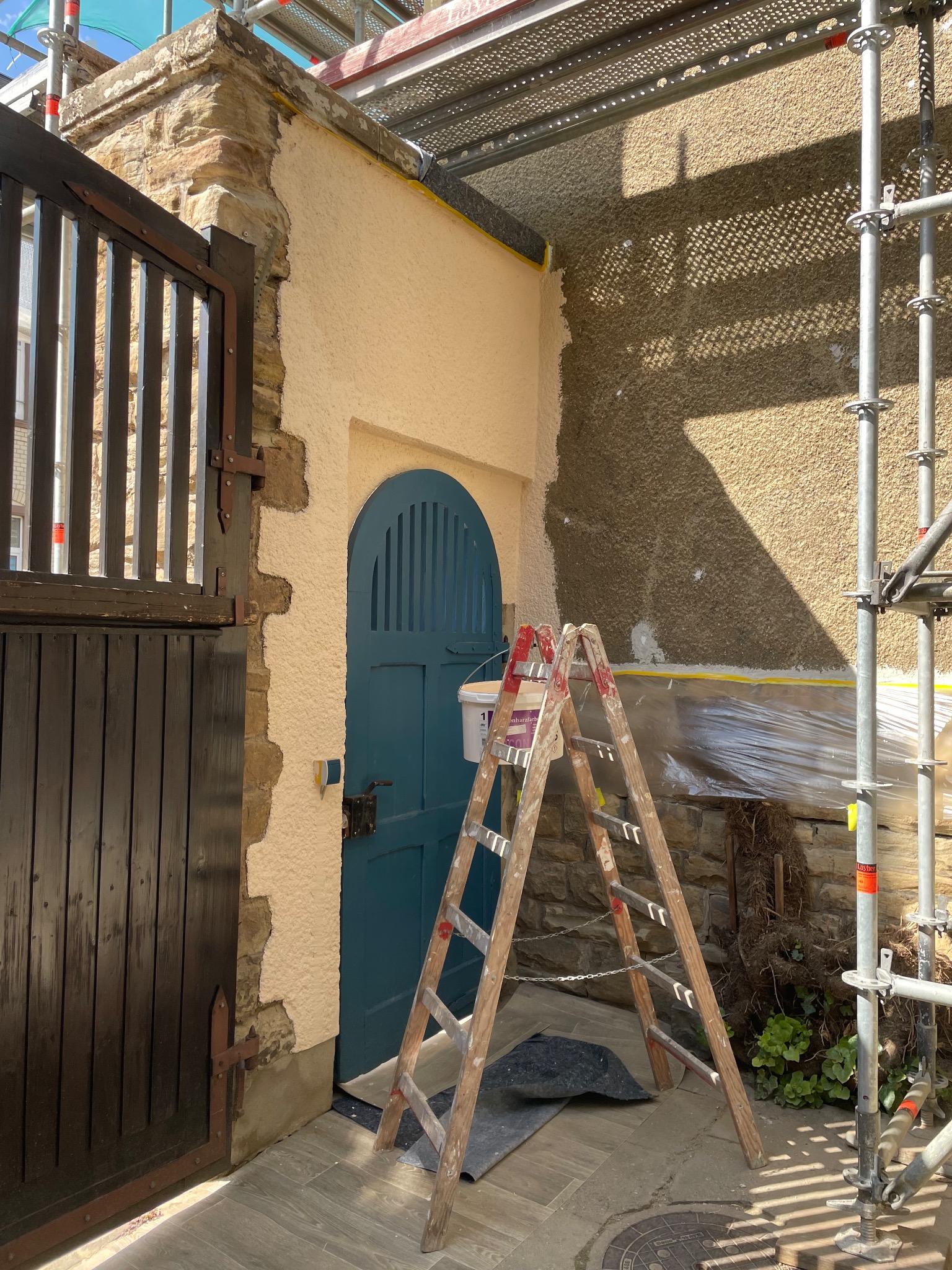 Elewacja budynku w trakcie prac: fragment otynkowanej ściany, drabina malarska, rusztowanie. Widoczna brama i drzwi. Prace prowadzone na zewnątrz budynku.
