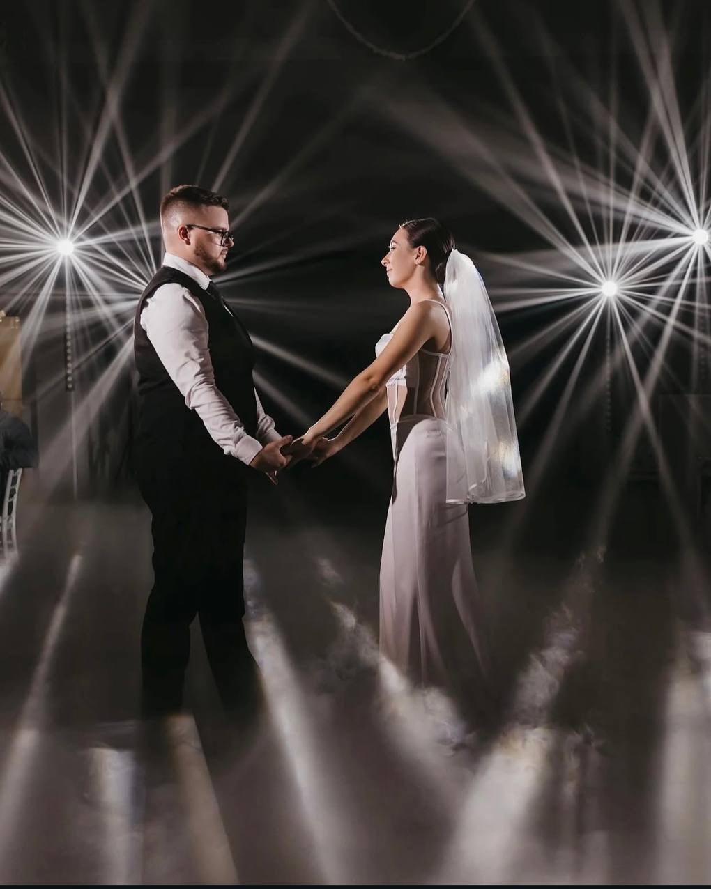 Para młoda trzymająca się za ręce w Białymstoku, oświetlona reflektorami podczas weselnego tańca w chmurach. Romantyczna atmosfera.