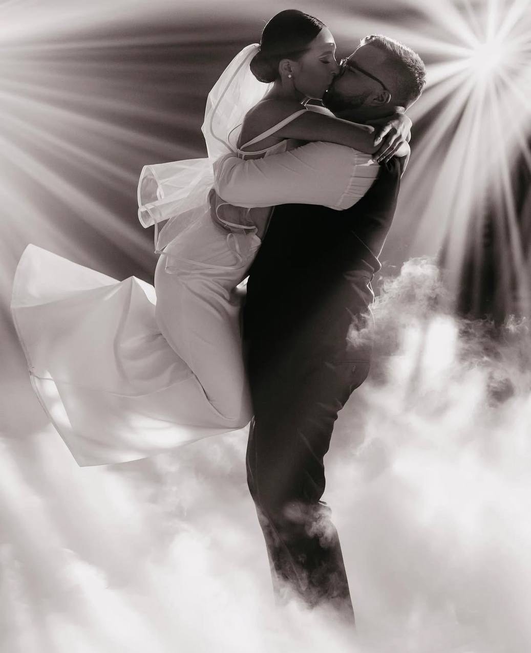 Czarno-białe zdjęcie nowożeńców: pan młody trzyma pannę młodą w objęciach, pocałunek na tle efektu mgły i promieni światła, tworząc romantyczną atmosferę.