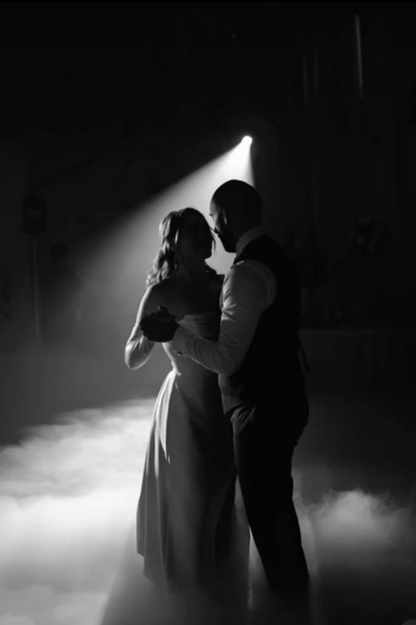 Czarno-białe zdjęcie pary młodej tańczącej w dymie, oświetlonej pojedynczym reflektorem podczas wesela. Sylwetki, intymna atmosfera, uroczystość.
