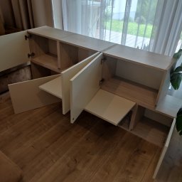 PARLAN Bartłomiej Giefert - Jasna, modułowa szafka RTV z otwartymi i zamkniętymi półkami, z widocznymi zawiasami, na tle okna z zasłonami i parkietu. Ujęcie z perspektywy podłogi.