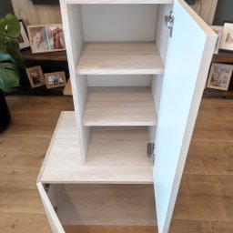 PARLAN Bartłomiej Giefert - Jasna szafka z otwartymi białymi drzwiczkami, widoczne półki wewnątrz. Na blacie zdjęcia w ramkach. Całość na tle drewnianej podłogi.