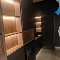 Nowoczesna łazienka z szafkami na wymiar z podświetleniem LED, czarnymi ścianami i pralką. Umywalka nablatowa i półki wnękowe dodają elegancji.