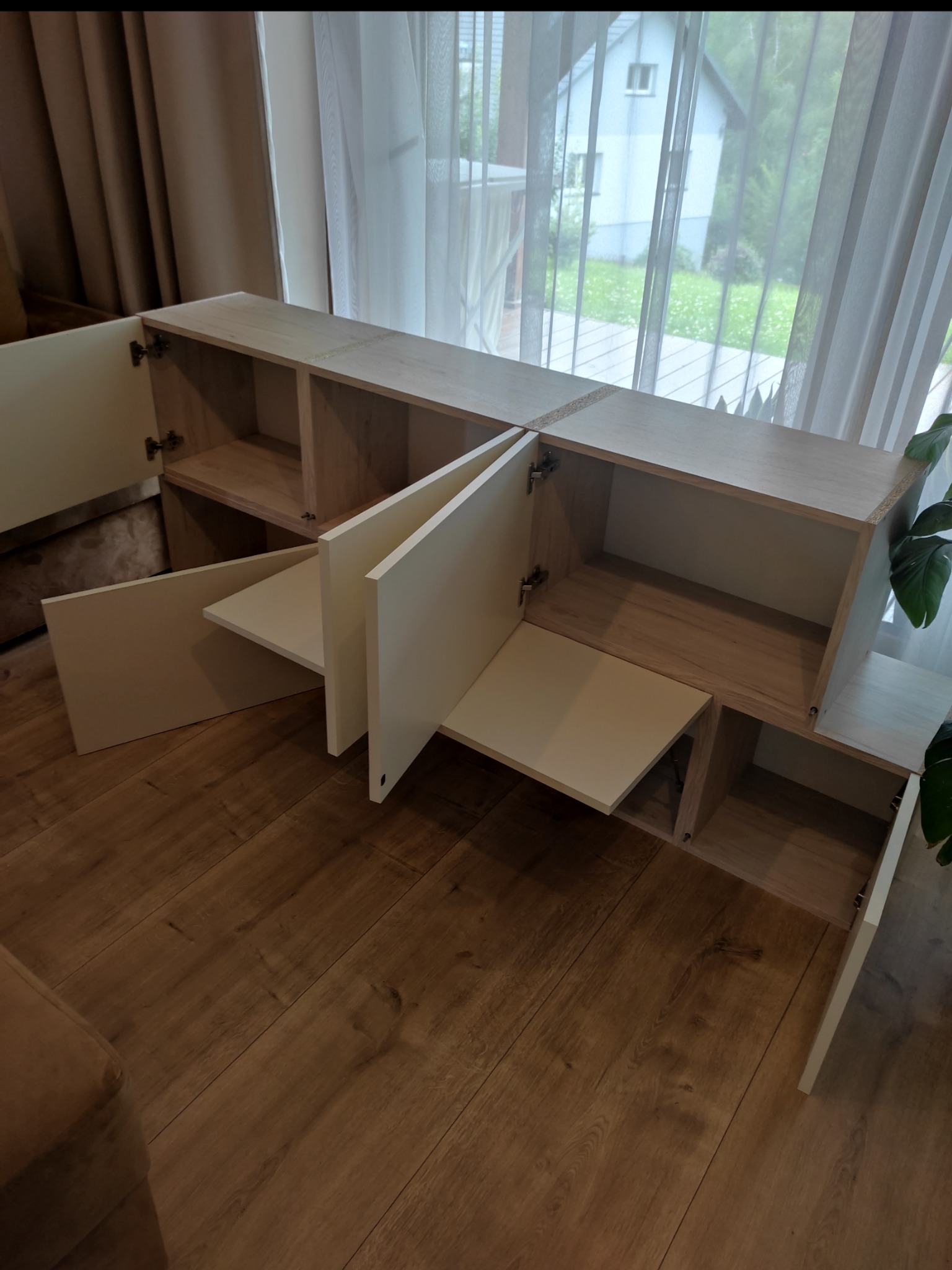 Jasna, modułowa szafka RTV z otwartymi i zamkniętymi półkami, z widocznymi zawiasami, na tle okna z zasłonami i parkietu. Ujęcie z perspektywy podłogi.