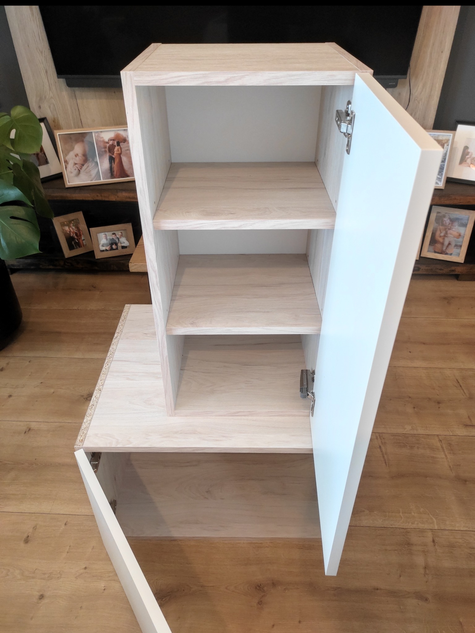 Jasna szafka z otwartymi białymi drzwiczkami, widoczne półki wewnątrz. Na blacie zdjęcia w ramkach. Całość na tle drewnianej podłogi.