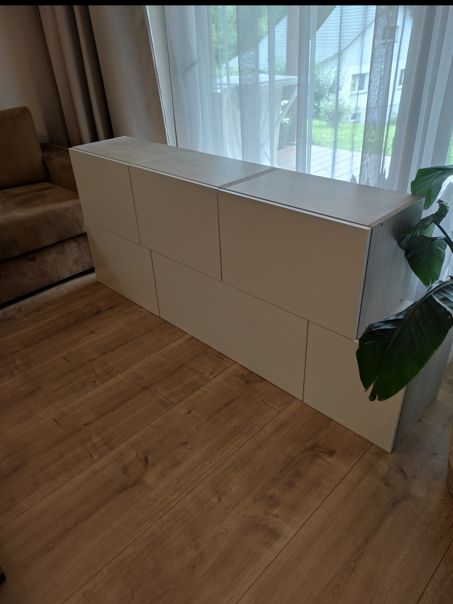 Biała szafka RTV z asymetrycznym układem frontów na drewnianej podłodze, w tle okno z firanką i zielony ogród. Minimalistyczny design.