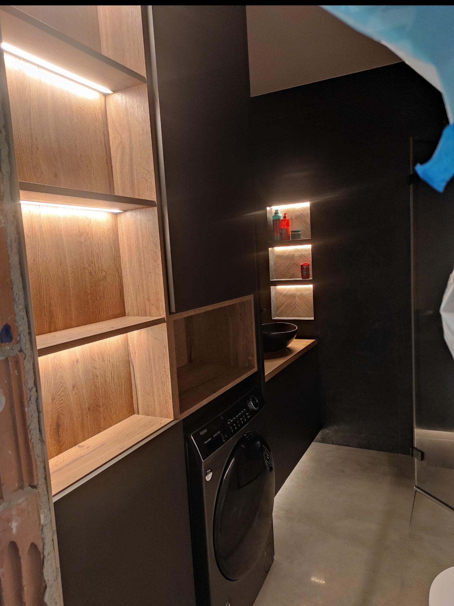 Nowoczesna łazienka z szafkami na wymiar z podświetleniem LED, czarnymi ścianami i pralką. Umywalka nablatowa i półki wnękowe dodają elegancji.