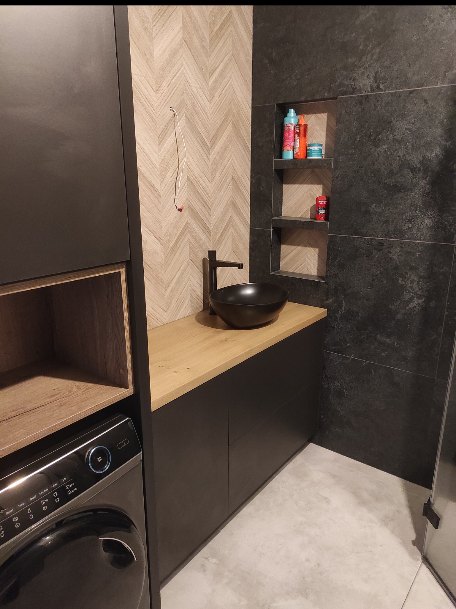 Nowoczesna łazienka z czarną umywalką nablatową, drewnianym blatem i szafkami w zabudowie. Na ścianie dekoracyjne płytki w jodełkę i wnęka z kosmetykami.