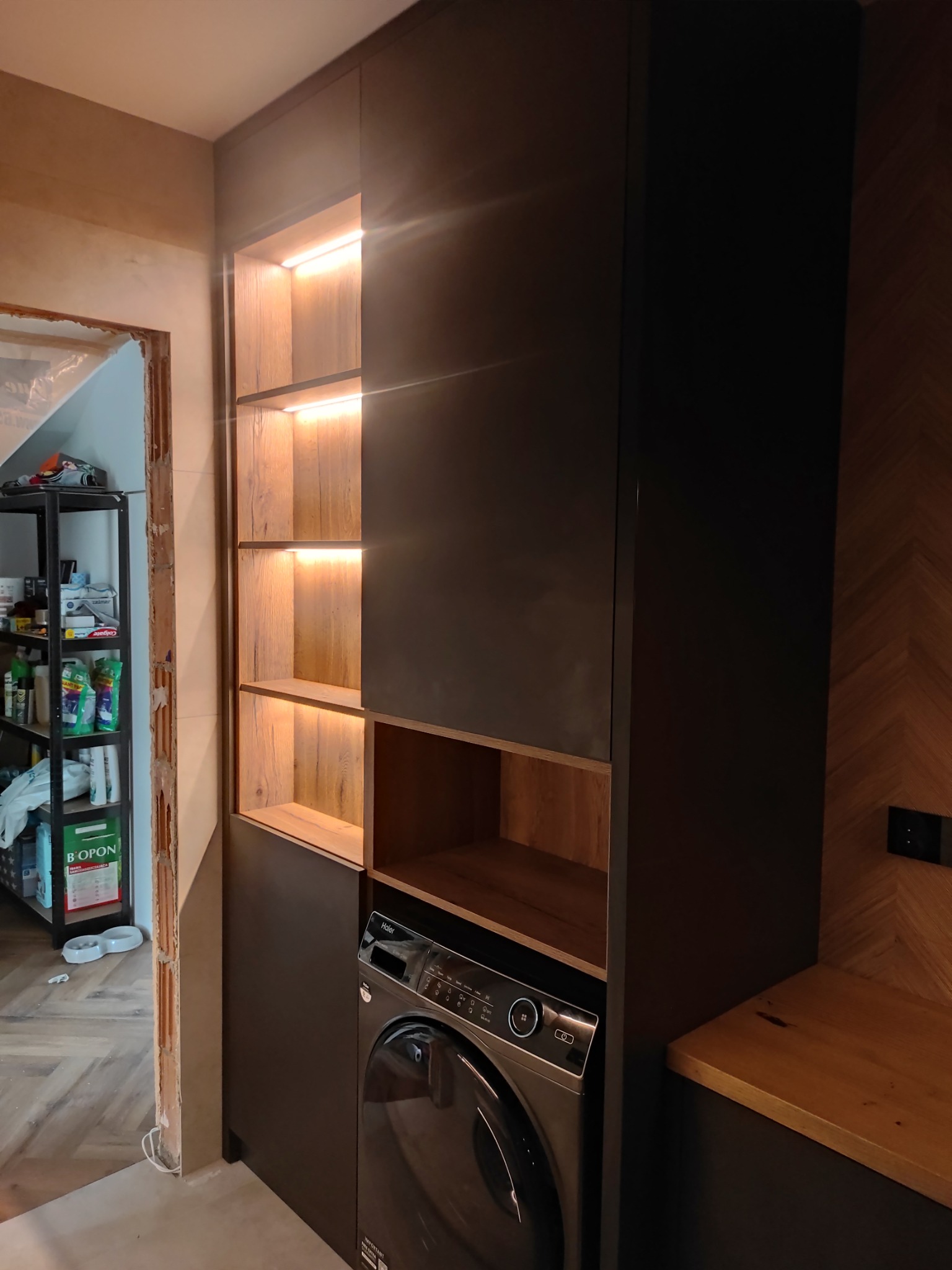 Ciemna szafa na wymiar z oświetleniem LED i drewnianymi półkami, obok pralka. Na ścianie dekoracyjna boazeria w jodełkę. Widoczne niedokończone wykończenie ściany.