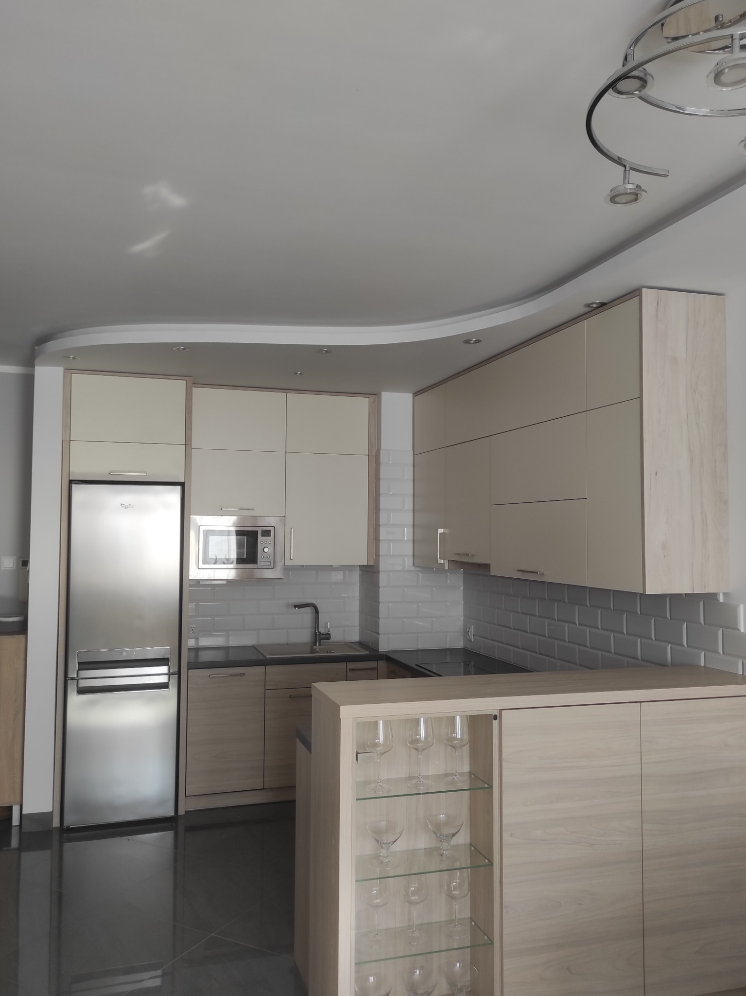 Jasna kuchnia z beżowymi frontami i szarą podłogą. Na wyspie szklane półki z kieliszkami. Białe płytki na ścianie i nowoczesne oświetlenie sufitowe.