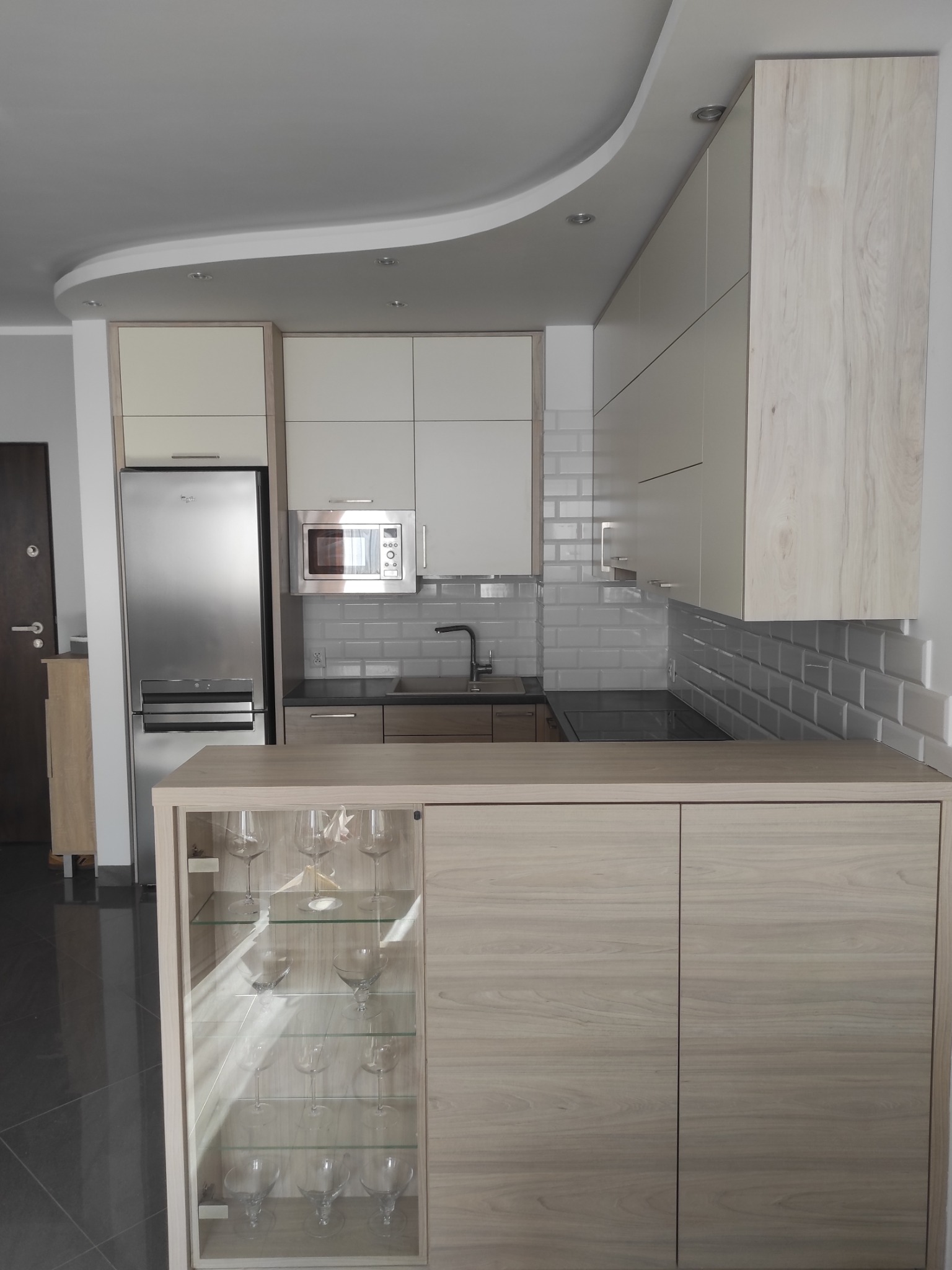 Jasna, nowoczesna kuchnia z drewnianymi frontami, szklaną witryną i srebrnym sprzętem AGD. Widoczne białe płytki na ścianie i dekoracyjny sufit.