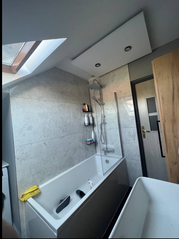 Wykończona łazienka na poddaszu z wanną, szklanym parawanem i płytkami imitującymi marmur. Widoczne okno dachowe i oświetlenie punktowe.