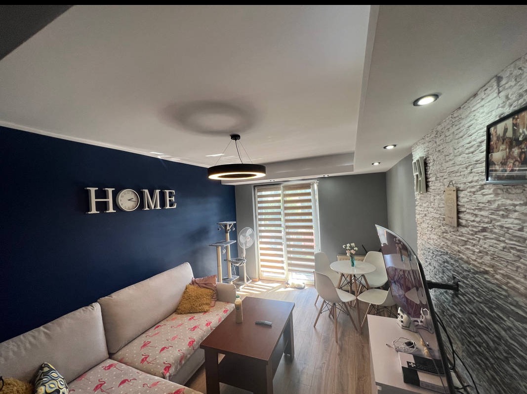 Nowoczesny salon po remoncie: ciemnoniebieska ściana z napisem 'HOME', dekoracyjna ściana z kamienia, okrągła lampa, parkiet. Widok na jadalnię i okno.