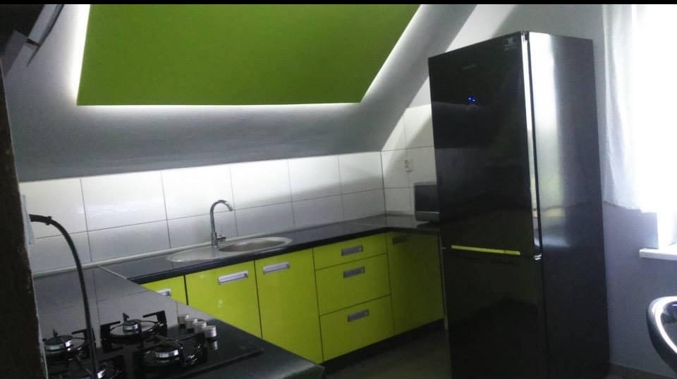 Nowoczesna kuchnia na poddaszu: limonkowy front mebli, czarny blat i lodówka, białe płytki, oświetlenie LED. Ukośny sufit i okno dodają unikalnego charakteru.