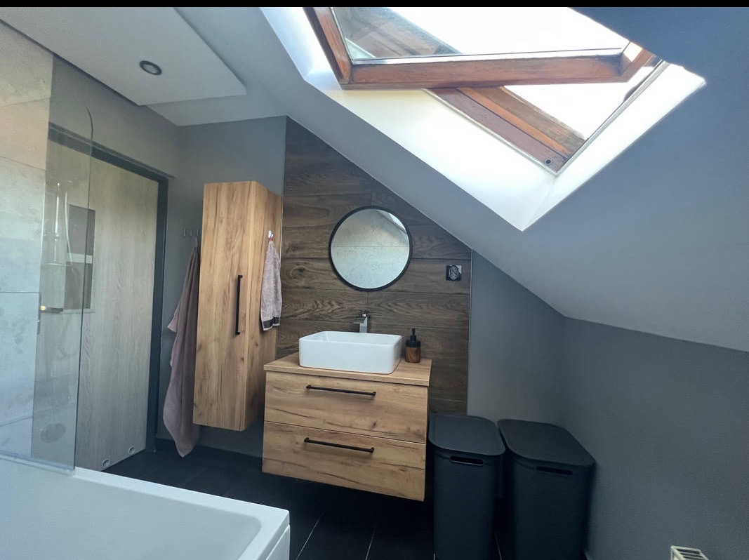 Nowoczesna łazienka na poddaszu ze skośnym oknem dachowym. Umywalka nablatowa, szafka z drewnianym dekorem, czarne pojemniki na pranie, szklany prysznic.