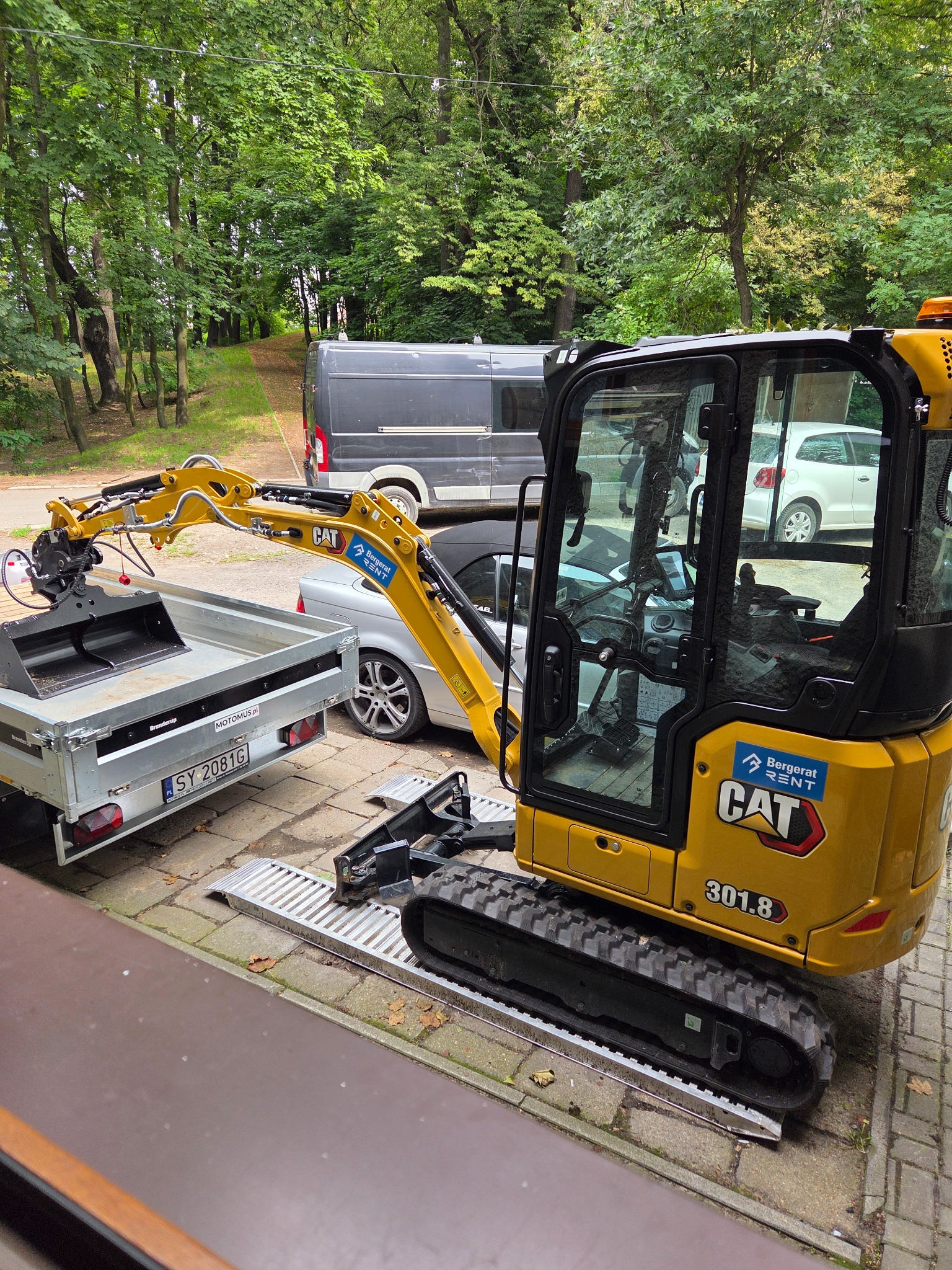 Żółta mini koparka CAT 301.8 na chodniku, z łyżką skierowaną w stronę przyczepy. W tle zieleń drzew i zaparkowane auta. Widoczna rampa najazdowa.
