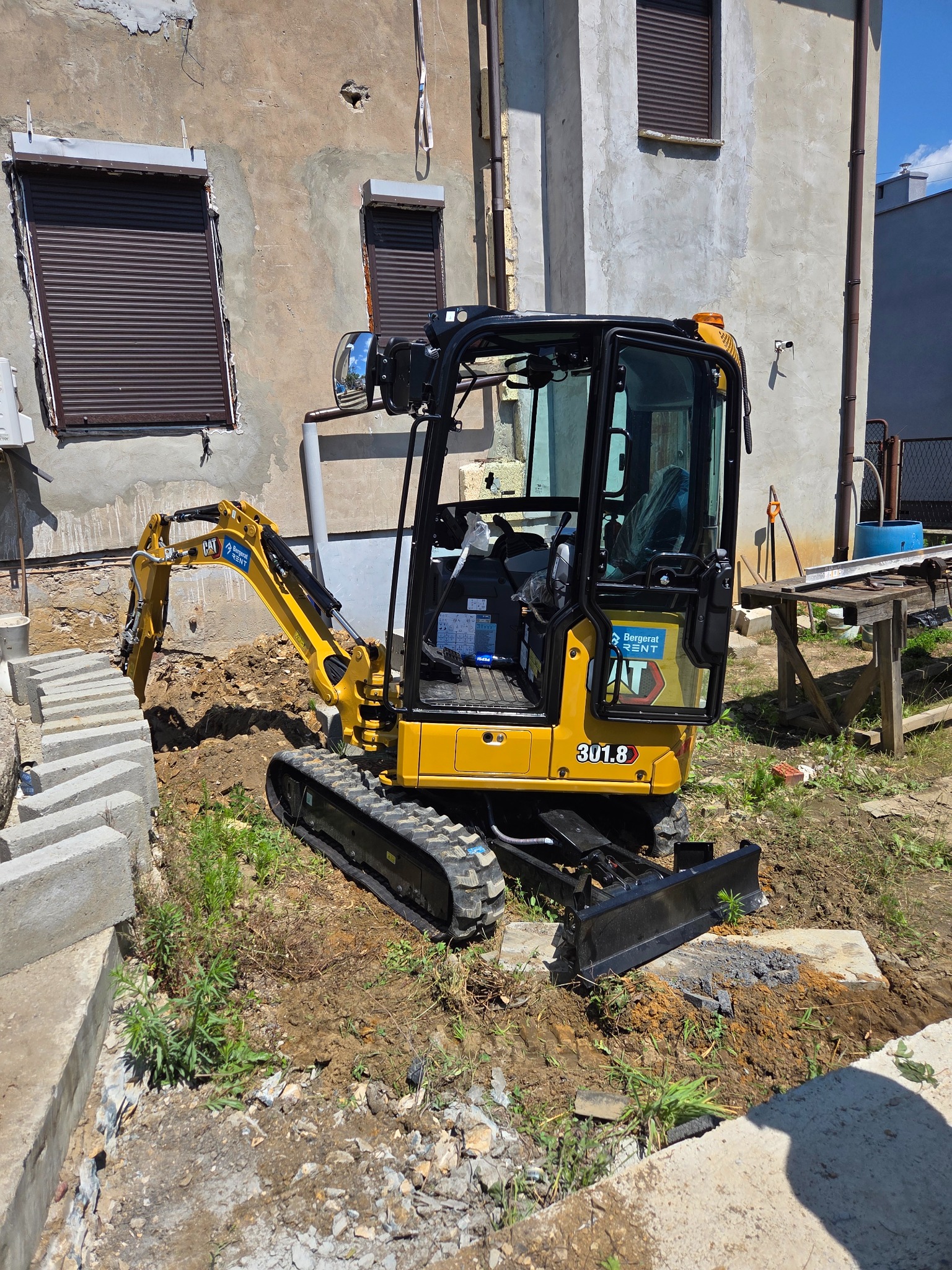 Żółta mini koparka CAT 301.8 na nierównym terenie z wykopem, obok budynku z zamkniętymi okiennicami. Widoczne betonowe elementy i narzędzia.