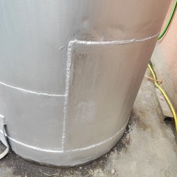 Szymon Kominkiewicz - Szczegół srebrnej, metalowej konstrukcji cylindrycznej z widocznymi spawami, stojącej na betonowej powierzchni. Widoczny fragment przewodu elektrycznego.