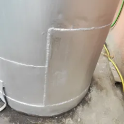 Szczegół srebrnej, metalowej konstrukcji cylindrycznej z widocznymi spawami, stojącej na betonowej powierzchni. Widoczny fragment przewodu elektrycznego.