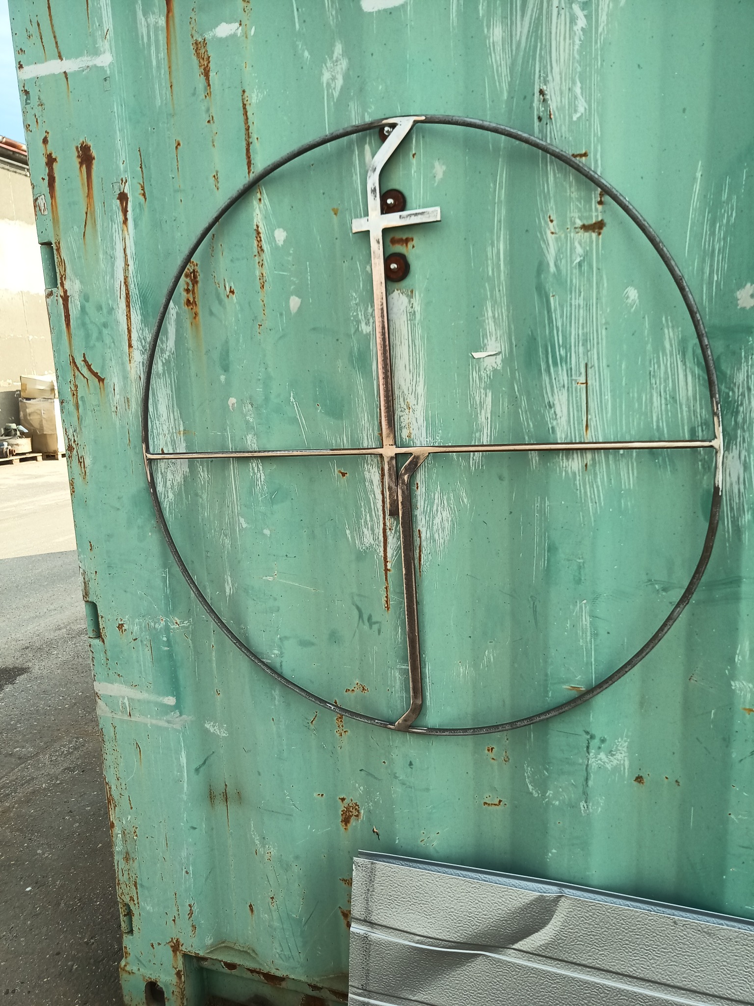 Metalowy okrąg z krzyżem przymocowany do zielonej, zardzewiałej ściany. Konstrukcja ślusarska w surowym stanie, widoczne elementy mocujące i spawane. Detal wykonania.