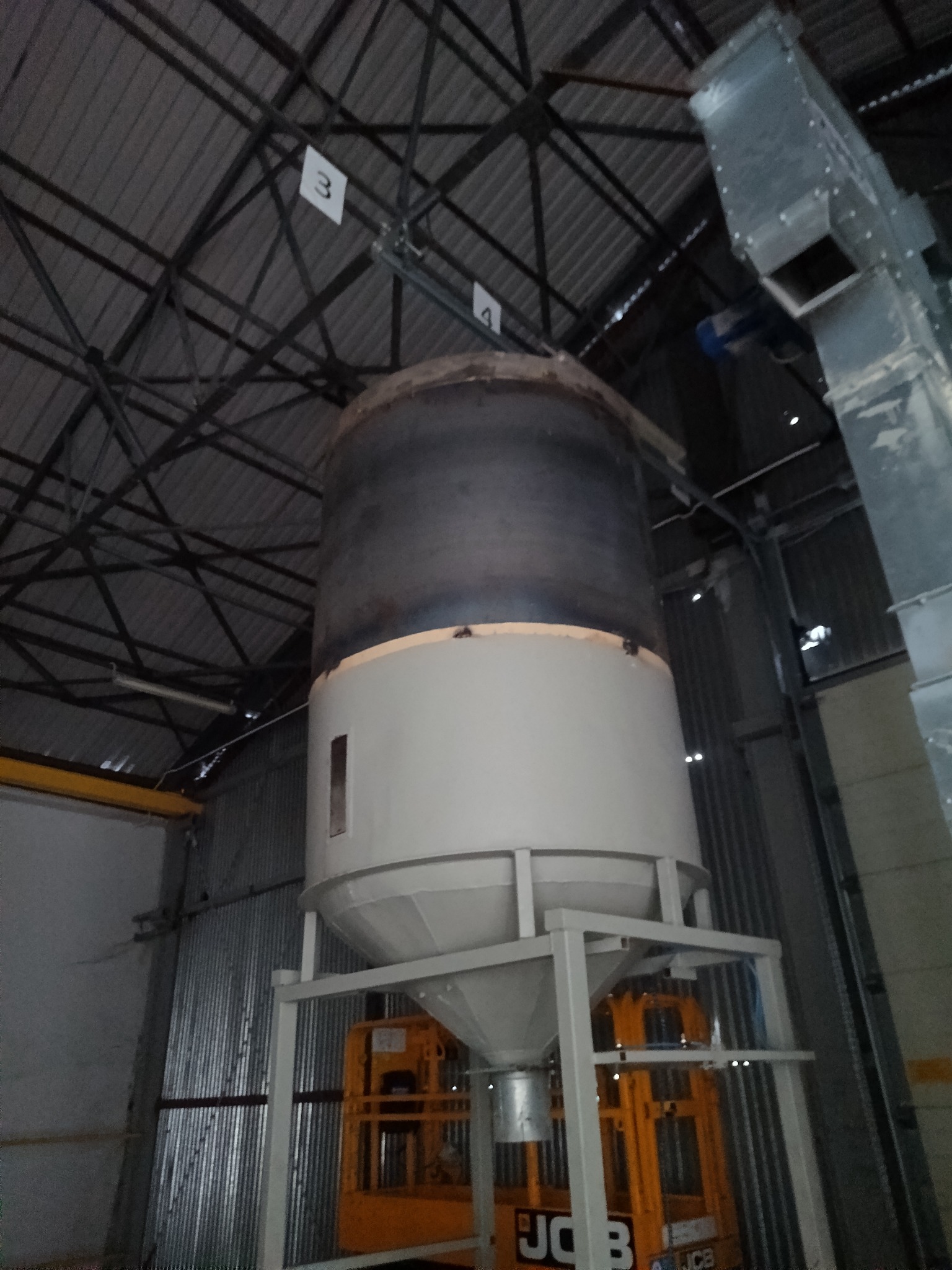 Wnętrze hali z metalową konstrukcją wsporczą dla zbiornika, widoczny podnośnik JCB. Górna część zbiornika ciemna, dolna biała. Oświetlenie halogenowe.