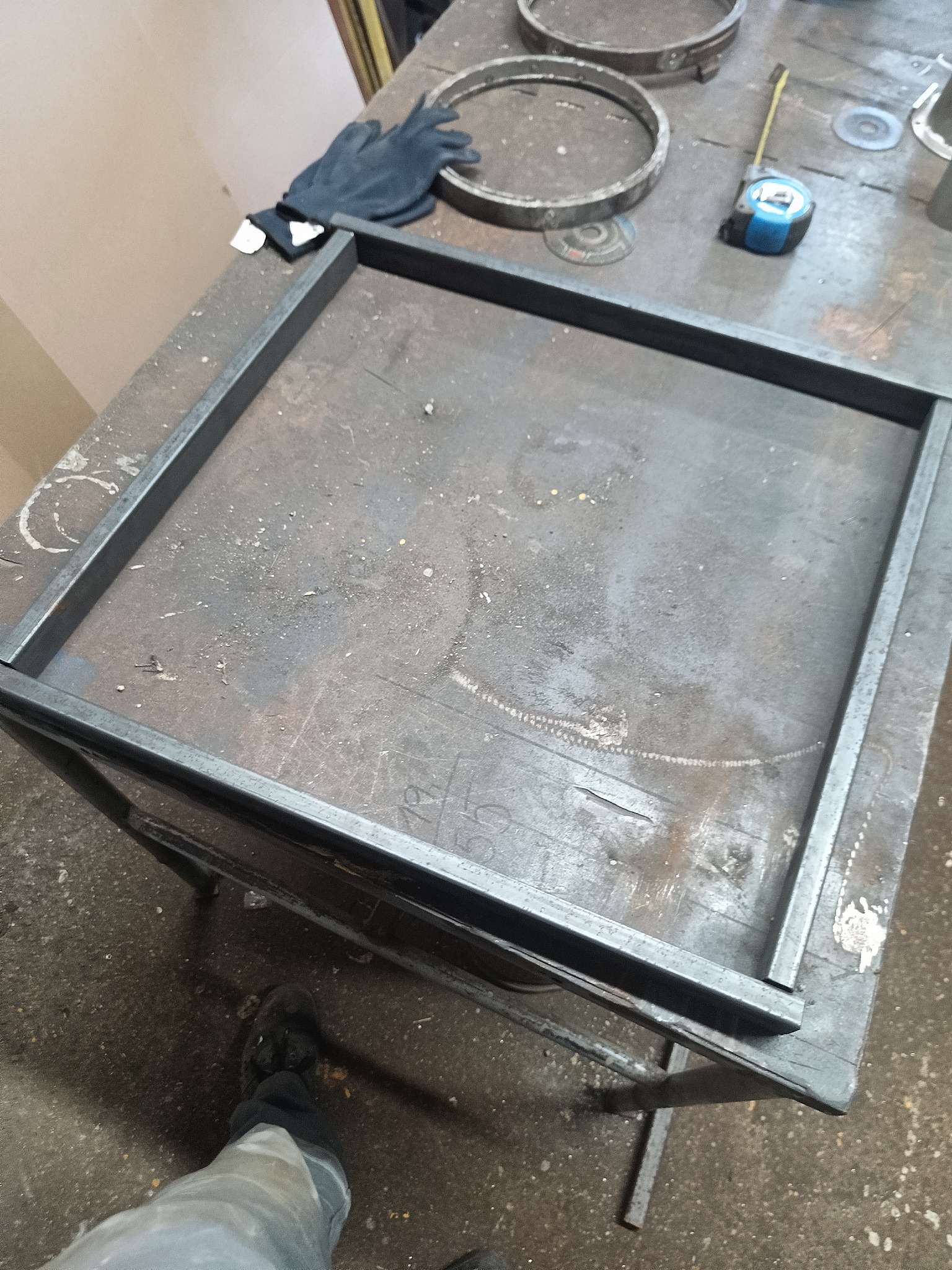 Spawana, kwadratowa rama z metalu na stole warsztatowym w Pyzdrach. Obok leżą rękawice robocze i miara zwijana. Widoczne ślady pracy ślusarskiej.