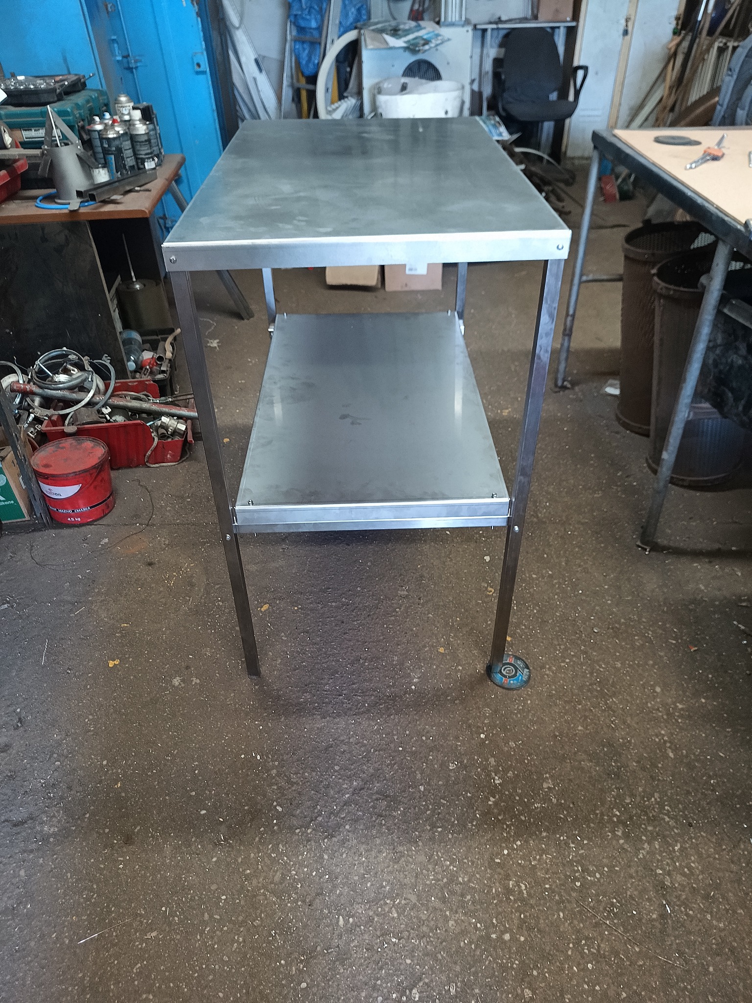 Stalowy stół warsztatowy z dwoma poziomami, spawany, z blachy, na tle warsztatu. Widoczne ślady użytkowania, co dodaje autentyczności.