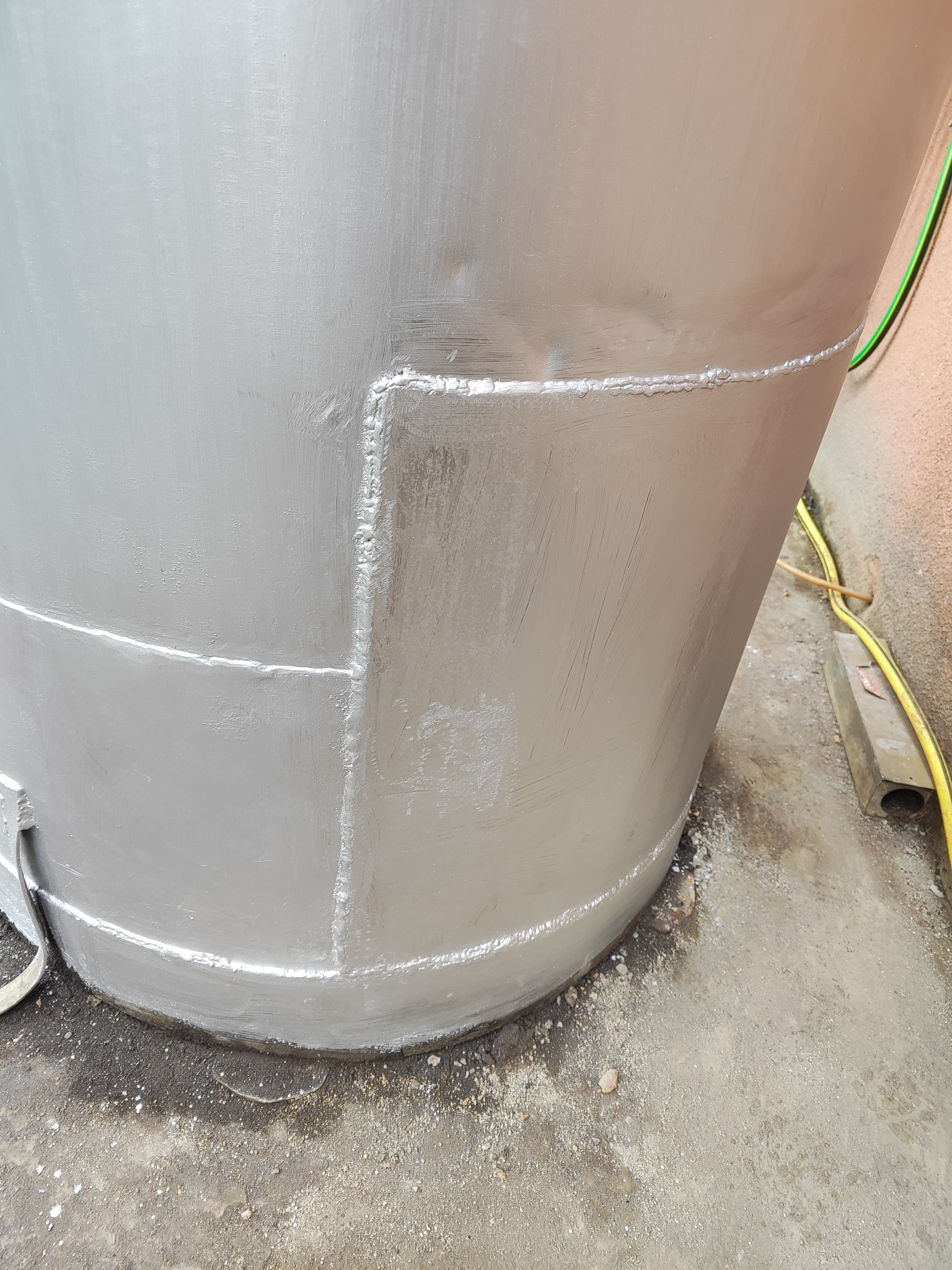 Szczegół srebrnej, metalowej konstrukcji cylindrycznej z widocznymi spawami, stojącej na betonowej powierzchni. Widoczny fragment przewodu elektrycznego.