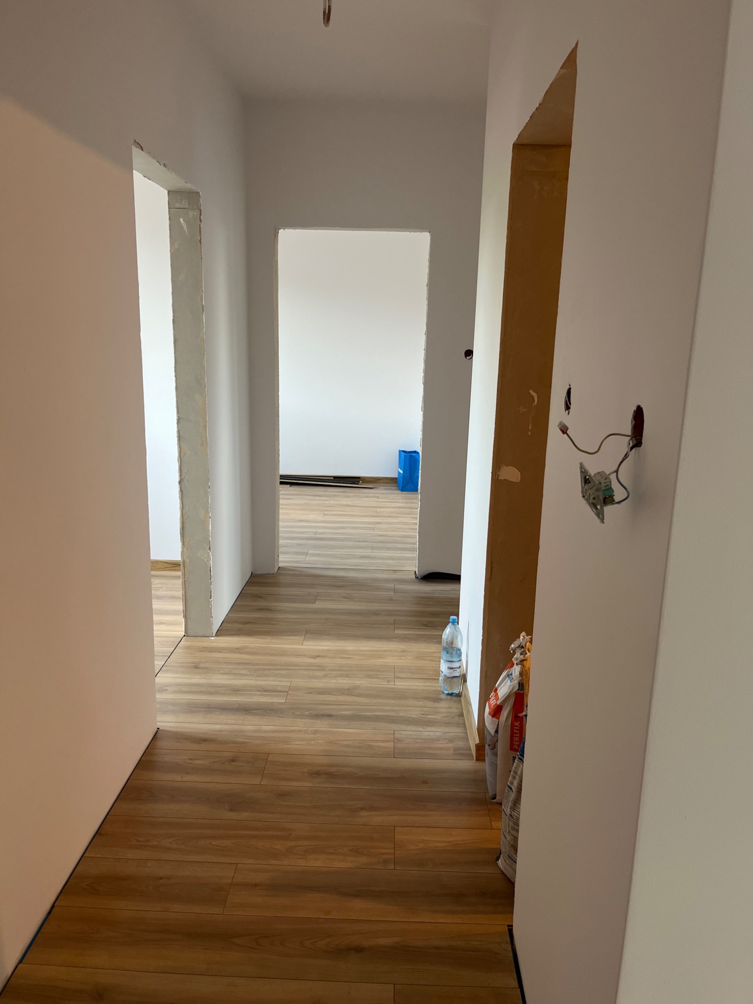 Wnętrze w trakcie wykończenia: panele podłogowe, świeżo pomalowane ściany z widocznymi przewodami elektrycznymi, torby z klejem i butelka wody.