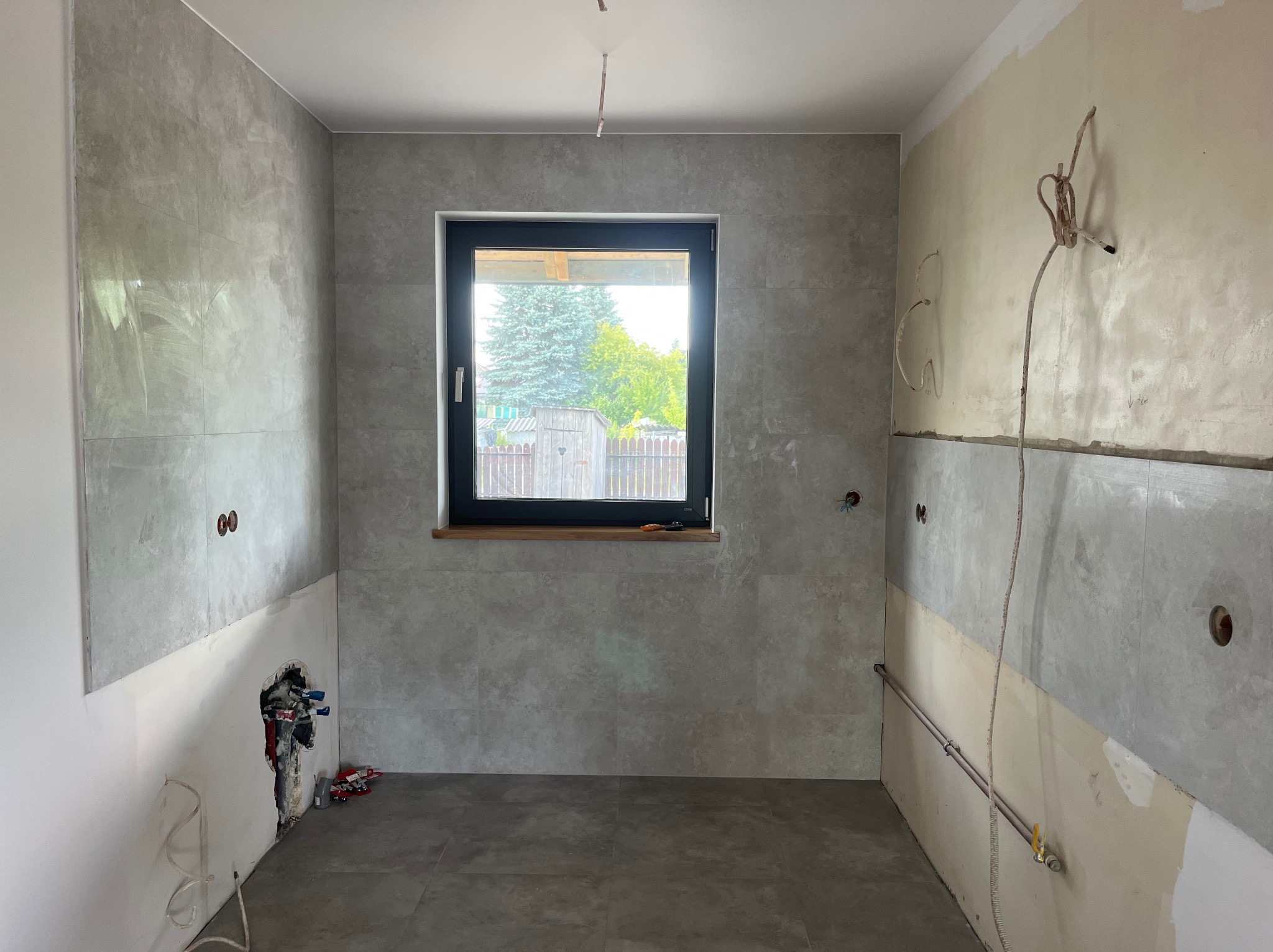 Szara, częściowo wykończona łazienka z oknem. Widoczne płytki na ścianach, instalacje, fragmenty ścian bez wykończenia. Prace w toku.