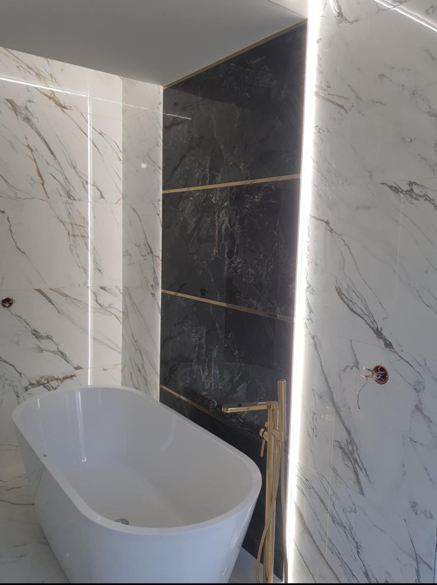 Elegancka łazienka z wolnostojącą wanną, biało-czarnymi płytkami imitującymi marmur, złotą armaturą i subtelnym oświetleniem LED. Widoczne detale wykończenia.