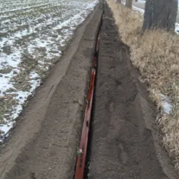 Wykopany rów w ziemi z pomarańczową rurą kablową w środku, widok z góry. Po lewej pole z resztkami śniegu, po prawej trawa i drzewa. Pochmurny dzień.