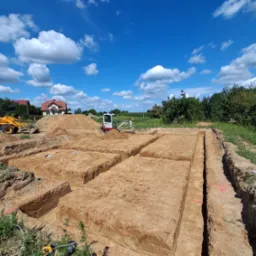 Wykopane rowy pod fundamenty na działce, z widocznymi maszynami budowlanymi i błękitnym niebem z chmurami w tle. Prace ziemne w toku, przygotowanie do budowy.