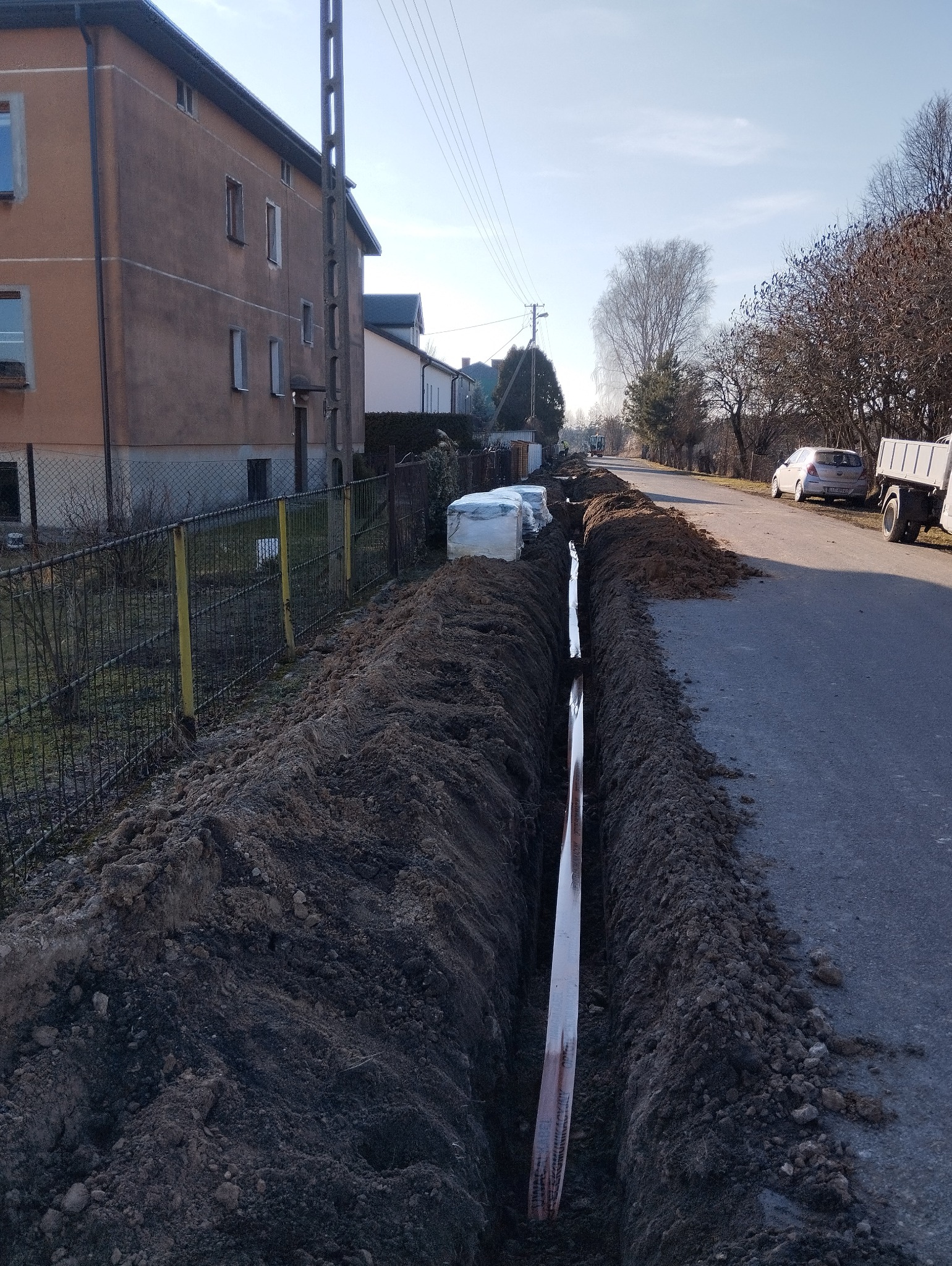 Wykopany rów wzdłuż drogi, wewnątrz rury. Po bokach pryzmy ziemi, w tle budynki mieszkalne i zaparkowane pojazdy. Prace ziemne.