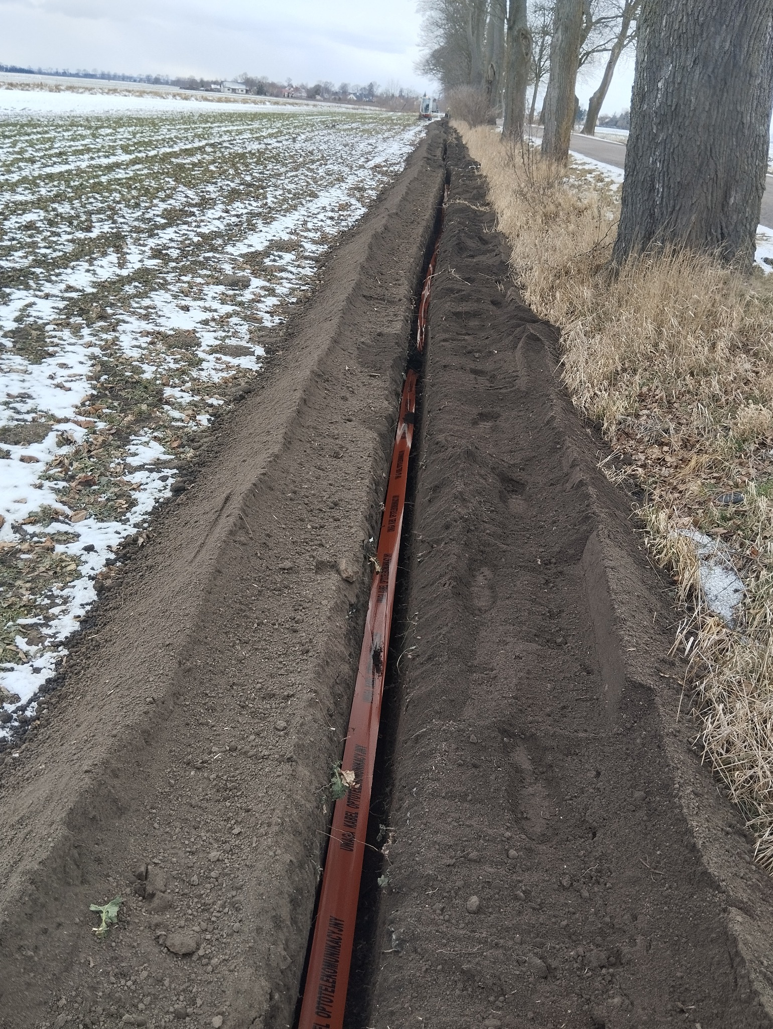 Wykopany rów w ziemi z pomarańczową rurą kablową w środku, widok z góry. Po lewej pole z resztkami śniegu, po prawej trawa i drzewa. Pochmurny dzień.