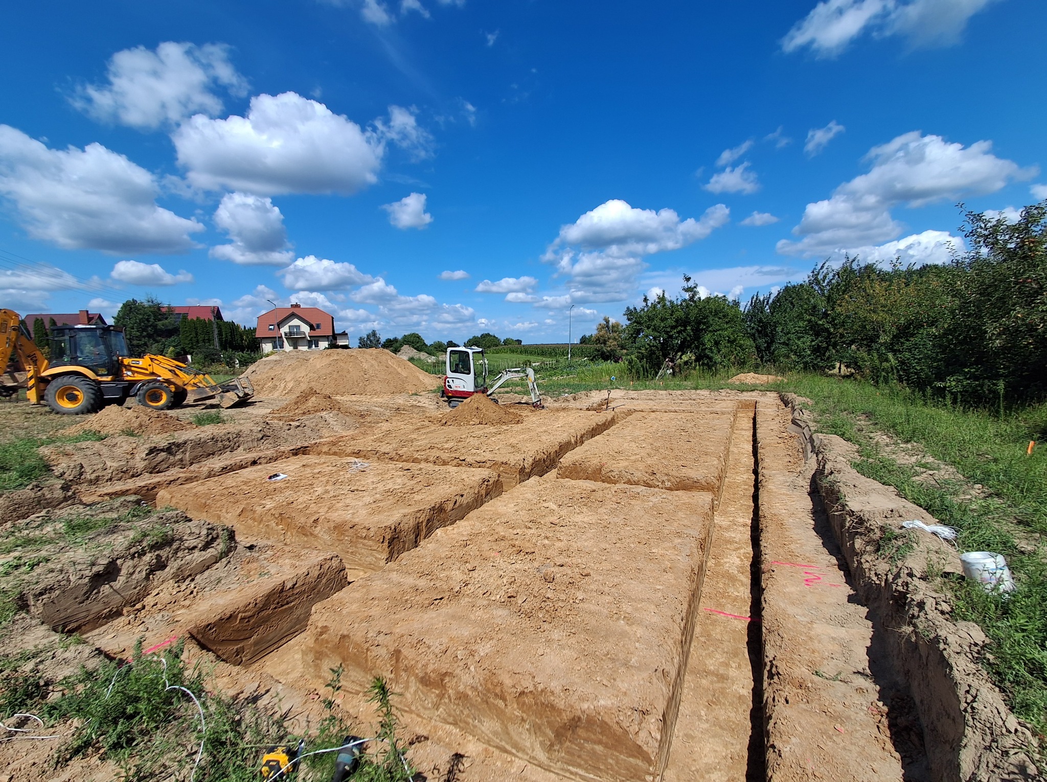 Wykopane rowy pod fundamenty na działce, z widocznymi maszynami budowlanymi i błękitnym niebem z chmurami w tle. Prace ziemne w toku, przygotowanie do budowy.