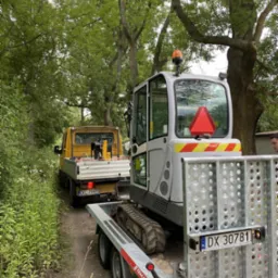 Transport minikoparki na przyczepie za żółtym samochodem dostawczym na leśnej drodze. Widoczne drzewa i zieleń w tle.