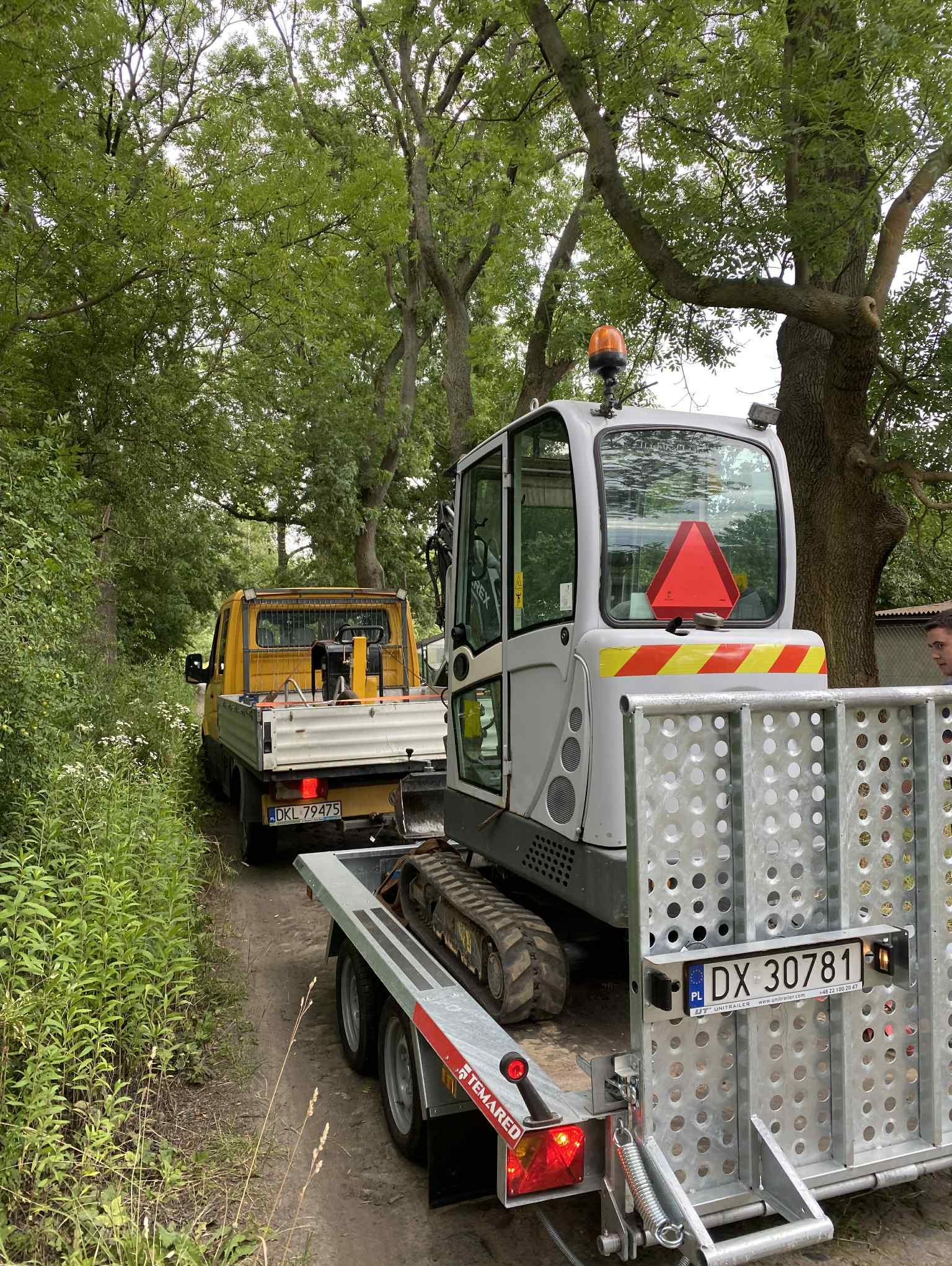 Transport minikoparki na przyczepie za żółtym samochodem dostawczym na leśnej drodze. Widoczne drzewa i zieleń w tle.