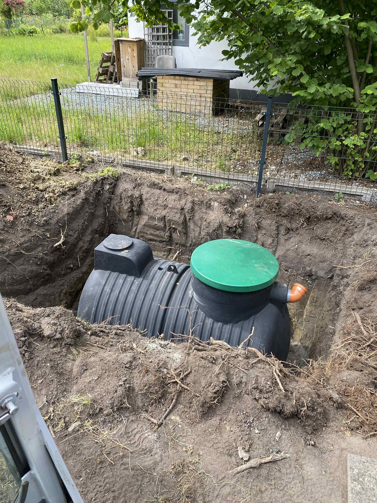 Czarny zbiornik z zieloną pokrywą w wykopie ziemnym, widoczne korzenie i fragment ogrodzenia panelowego w tle, sugerujący prace instalacyjne.