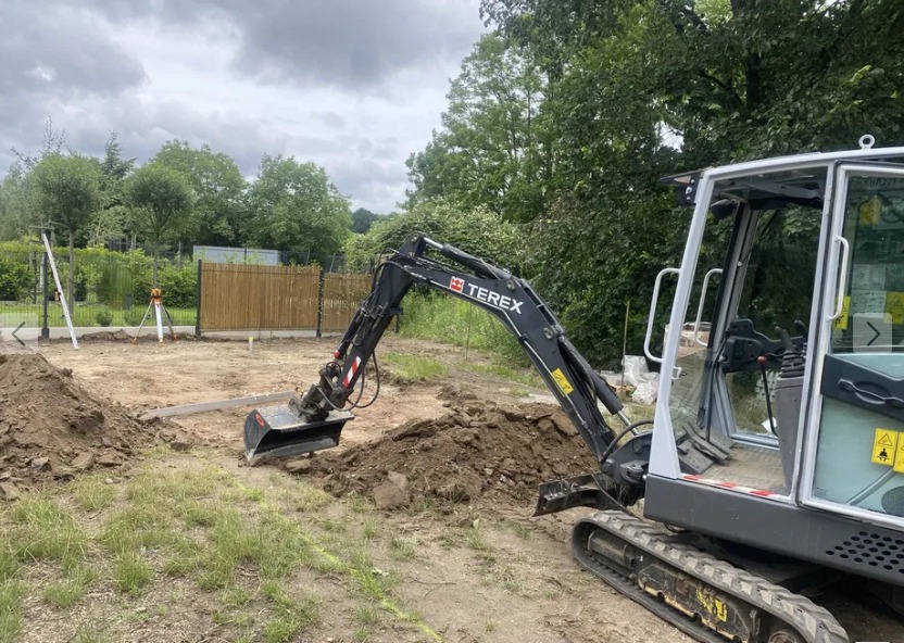 Minikoparka Terex przy wykopie, widoczny fragment ogrodzenia panelowego i niwelator. Prace ziemne w toku, teren przygotowywany pod inwestycję.