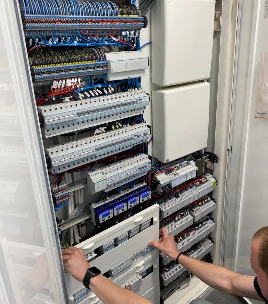 Otwarta szafa elektryczna z widocznymi bezpiecznikami, przewodami i elementami sterującymi. Elektryk w trakcie montażu panelu, widoczne dłonie i zegarek.