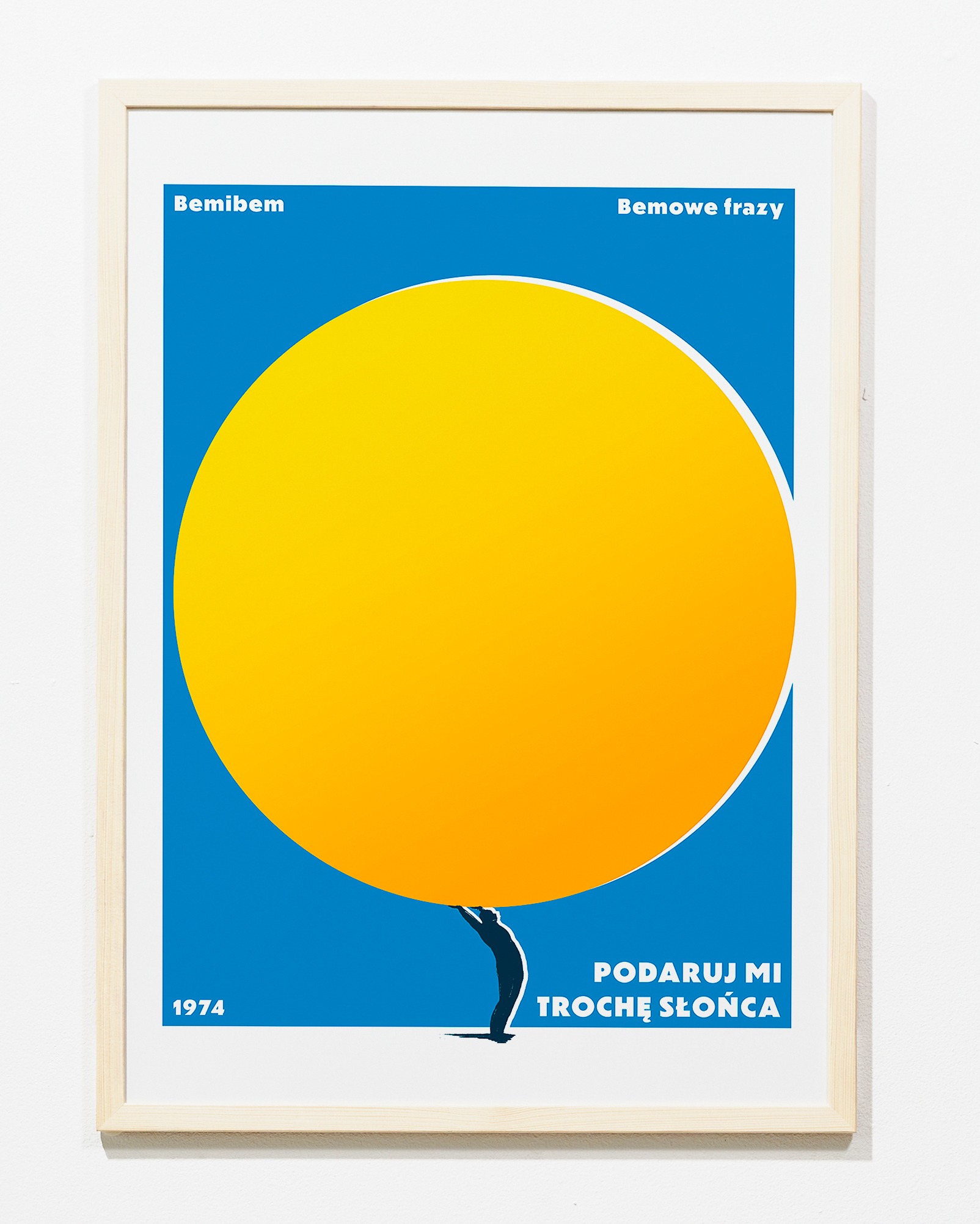 Oprawiony plakat z 1974 roku w stylu retro: na niebieskim tle żółte słońce dźwigane przez sylwetkę człowieka. Napisy 'Bemibem', 'Bemowe frazy' i 'Podaruj mi trochę słońca'.