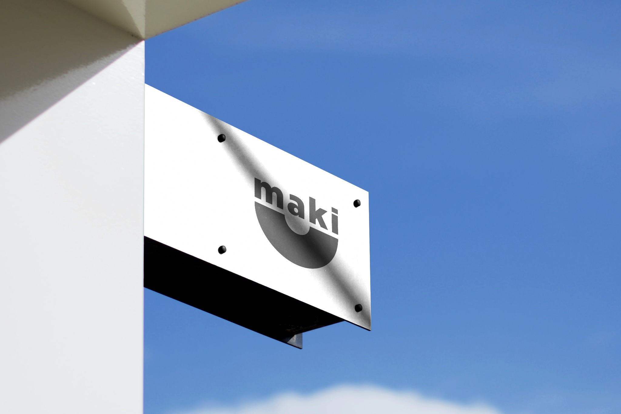 Biały szyld reklamowy z czarnym logo 'maki' na tle błękitnego nieba, zamocowany do rogu budynku. Widoczny fragment elewacji i mocowania szyldu.
