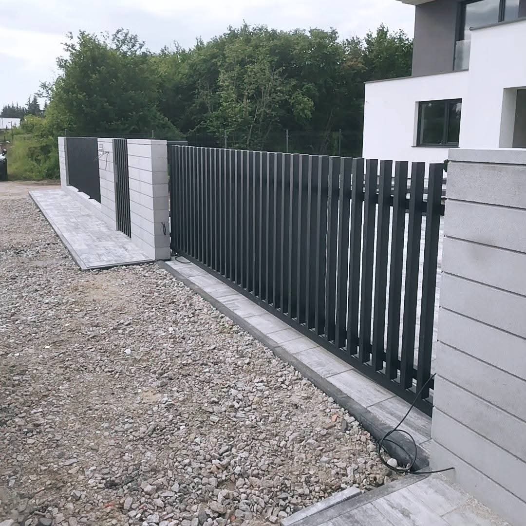 Nowoczesne ogrodzenie posesji z metalowymi sztachetami w kolorze antracytu i betonowymi słupkami. Widoczny kabel elektryczny do automatyki bramy.