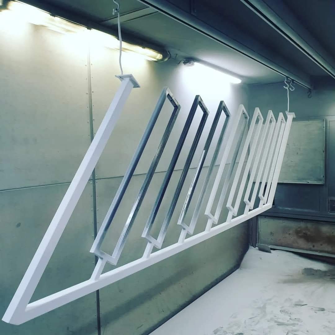 Metalowa konstrukcja z prostokątnymi ramami, częściowo pomalowana na biało, zawieszona w lakierni. Widoczne haki, oświetlenie i ślady farby na podłodze.