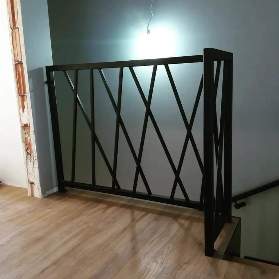 Czarna, metalowa balustrada schodowa z geometrycznym wzorem krzyżujących się prętów, zamontowana przy drewnianych schodach w nowoczesnym wnętrzu.