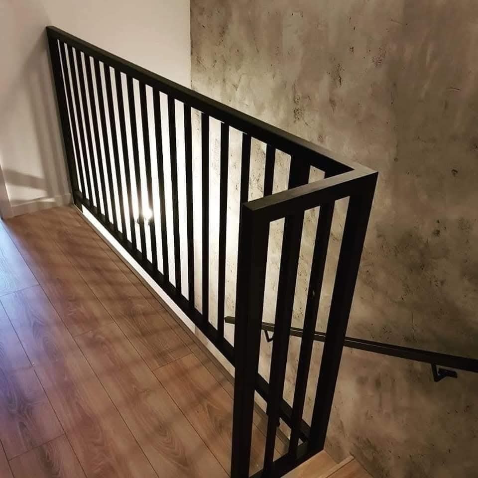 Nowoczesna, czarna balustrada schodowa z pionowymi szczebelkami, zamontowana przy ścianie o surowym, betonowym wykończeniu i drewnianej podłodze.